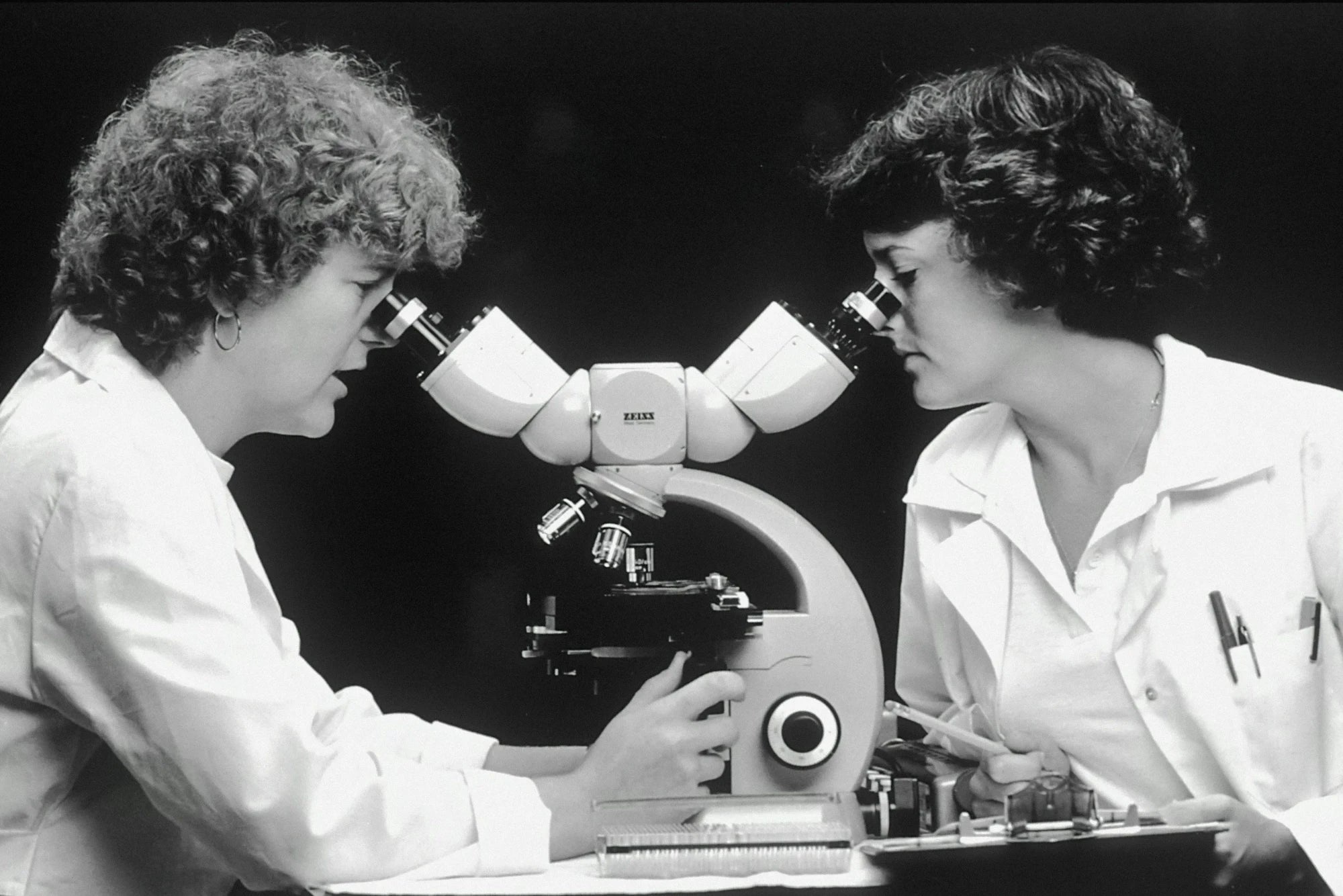 Two researchers in lab coats look into a shared microscope.